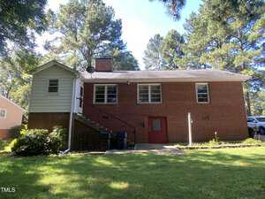A brick house with a side staircase and red door, surrounded by a grassy yard and trees. The house has a chimney and several windows, set against a clear blue sky. Trash bins are placed near the stairs.