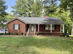 The single-story brick house at 303 Forest Road, Oxford, features a charming covered front porch with white columns and a small staircase leading to the front door. It's nestled among grass, shrubs, and tall trees under a partly cloudy sky, with a car parked on the left.