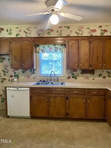 A vintage kitchen with wooden cabinets and floral wallpaper. A window with a patterned valance is above a double sink, and a dishwasher is to the left. A ceiling fan with a light is above, brightening the beige floor and countertops.
