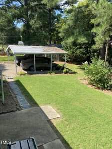 A backyard with a green lawn and a concrete path. A silver car is parked under a metal carport. Tall pine trees and shrubs surround the area, providing shade and privacy. A sunny day with clear skies.