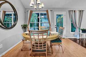 A dining room with a round wooden table and four chairs. A large circular mirror hangs on the wall. Windows with curtains bring in natural light. A potted plant sits on a side table, and a chandelier hangs above the table.