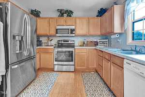 A modern kitchen with wooden cabinets, stainless steel appliances, and a double sink. A refrigerator is on the left, electric stove and microwave are centered, and a dishwasher sits below the counter on the right. Two textured rugs cover the floor.