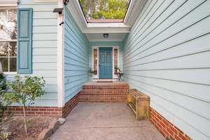 A cozy house entrance with light blue siding and a white trim. The doorway features a teal door adorned with a small decorative tree and flowers beside it. Brick steps lead to the door with a wooden bench along the right side.
