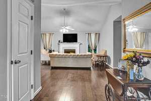 A cozy living room with a beige sofa facing a fireplace and a TV above it. Two chairs flank the fireplace. A decorative mirror and flowers adorn the wall on the right. Wooden floors and soft lighting create a warm atmosphere.