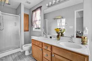 A bathroom with a glass shower, a wooden cabinet, and a large mirror above a double sink vanity. The vanity has a bouquet of flowers, a soap dispenser, and decorative candle holders. A window with a curtain provides natural light.