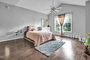 A spacious bedroom with a neatly made bed featuring blush-colored bedding and pink and gray pillows. There's a small side table and a dresser with a mirror. Large window with sheer curtains, wooden floor, ceiling fan, and decorative letters on the floor.