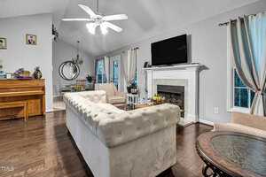 A cozy living room with a white tufted sofa facing a wall-mounted TV above a fireplace. A ceiling fan hangs above. A piano is on the left, and a dining area with a large mirror is visible in the background. Light curtains frame the windows.