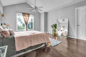 A cozy bedroom with a large bed covered in pink and green bedding, situated near a window with sheer curtains. There's a floor mirror reflecting a potted plant, and a light blue rug on the wooden floor. Ceiling fan overhead and soft natural light entering.