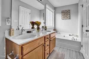 A bathroom features a double sink vanity with wooden cabinets, a large mirror, and a countertop with flowers and toiletries. A bathtub is on the right, beneath a window, with art on the wall. The floor is covered with gray tiles.