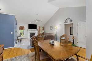 A cozy living and dining area with a vaulted ceiling. A wooden dining table with chairs is in the foreground. The living room features a sectional sofa, a fireplace, and a wall-mounted TV. Neutral walls and hardwood floors enhance the space.