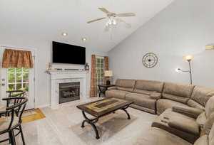 A cozy living room with a beige sectional sofa, a wooden coffee table, and a wall-mounted TV above a white fireplace. There's a ceiling fan, a decorative wall clock, and windows with brown curtains. Soft lighting and a neutral color palette create a warm atmosphere.