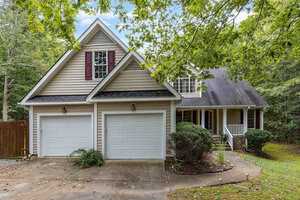 A two-story suburban house with beige siding and a dark roof, featuring a two-car garage. Surrounded by trees, it has a driveway and a small front porch with white railings. Lush greenery lines the pathway to the entrance.