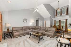 Spacious living room with beige sectional sofa, wooden coffee table, and bar stools. High ceiling with ceiling fan. Dining area in the background with large windows and curtains. Modern decor with a wall clock and carpeted floor.