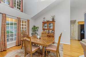 A dining room with a wooden table and four chairs on a patterned rug. A wooden hutch with decorative items is against the wall. Large windows with patterned curtains let in natural light. A plant is in one corner, and a hallway is visible.