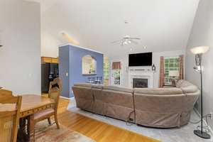 A living room with a large beige sectional sofa facing a fireplace and wall-mounted TV. The room has hardwood floors, a high ceiling with a fan, and a window with curtains. A dining table and chairs are visible to the left. The kitchen is in the background.
