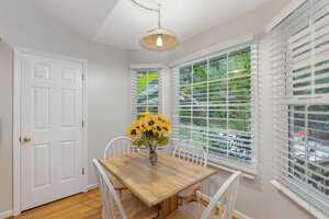 A cozy dining nook with a wooden table and four white chairs. A vase with sunflowers sits on the table. Large windows with blinds overlook a green, leafy garden. A pendant light hangs above the table, illuminating the space.