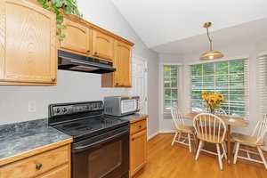 A kitchen with wooden cabinets, black countertops, and a black stove. A small dining table with four chairs is set by a window with blinds, adorned with a vase of sunflowers. A microwave sits on the counter, and a hanging light fixture is above the table.