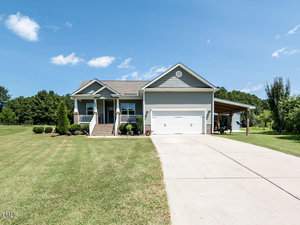 Nestled in Franklinton, 2630 Pauline Oaks Drive is a charming single-story home with light gray siding and a gabled roof. It boasts a front porch with stairs, a spacious driveway leading to a two-car garage, and a neatly manicured lawn. The sky is clear with just a few clouds.