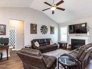 A cozy living room with two brown leather sofas, a fireplace with a TV above, and decorative wall art. A small desk with a computer sits in the corner. The room has wooden floors and a ceiling fan, with a glimpse into a bathroom.