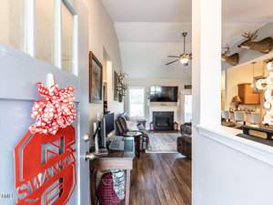 Open door revealing a cozy living room with hardwood floors, a fireplace, and a large TV. Mounted deer heads adorn the walls. A polka dot bow decorates the door, and a computer desk is visible to the left. Bright natural lighting fills the space.
