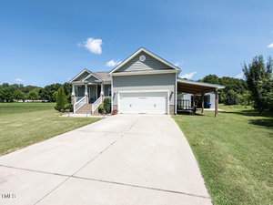 A light gray single-story house with a porch and a two-car garage sits on a spacious lawn. A paved driveway leads up to the house. A side carport is visible, and there are trees and a clear blue sky in the background.
