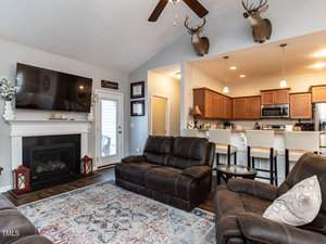 A cozy living room with a large TV above a white fireplace. Two brown sofas are arranged around a patterned rug. The kitchen area with wooden cabinets is visible, and two deer heads are mounted on the wall. A ceiling fan hangs overhead.