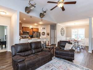 Open-concept living room and kitchen with brown sofas, wooden cabinets, and a dining table. Ceiling fan, mounted deer heads, and a large rug add decor. Brightly lit with natural light from windows.
