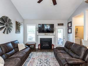 A cozy living room with two brown leather sofas facing a fireplace. A television is mounted above the white mantel. Two decorative lanterns sit by the fireplace, and a patterned rug covers the floor. The ceiling is high and vaulted.
