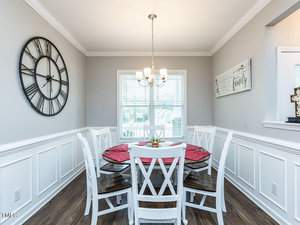 A dining room with a round table set with red placemats and white wooden chairs. A large clock decorates the gray wall, and a chandelier hangs from the ceiling. A window with blinds allows natural light in, and a "Family" sign is on the right wall.