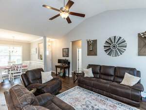 A cozy living room with brown leather sofas, decorative pillows, and a patterned rug. Wall decor includes a metal windmill piece. A ceiling fan hangs above. In the background, there's a dining area with a white table and chairs.