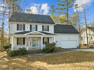 A two-story white house with a gray roof and a front porch, featuring an American flag. It has a large two-car garage, surrounded by trees with autumn foliage and a spacious lawn. A clear blue sky is in the background.