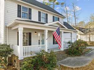 A two-story white house with a front porch, black shutters, and an American flag. It is surrounded by bushes, and there's a driveway on the right. The sky is clear and blue.