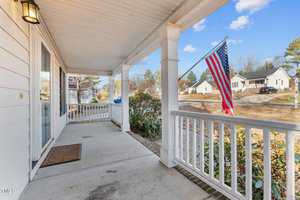 A suburban front porch with white railings and a welcome mat. An American flag is displayed on a post. The background shows a quiet street with houses and trees under a clear blue sky.
