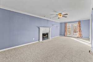A spacious living room with light blue walls features a white fireplace, beige carpet, a ceiling fan with lights, and French doors leading to an outdoor area with floral curtains. Shelving units are installed on the wall near the fireplace.