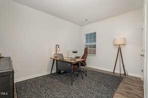 A minimalist home office with a wooden desk, laptop, and brown chair on a gray carpet. A tall floor lamp stands in the corner next to a window with blinds. The walls are plain white, and the flooring is a light wood finish.