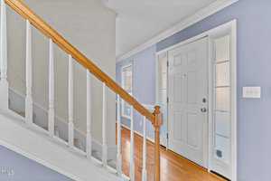 A hallway with light blue walls and wooden flooring. The white front door has glass panels on each side. A staircase with wooden railings and white spindles leads upward. Natural light brightens the space through a nearby window.