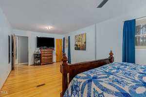 Bedroom with light blue walls, featuring a dark wooden bed with a blue and white patterned quilt. A dresser with a TV on top is against the far wall, and two windows with blue curtains let in natural light. Wood flooring adds warmth to the room.