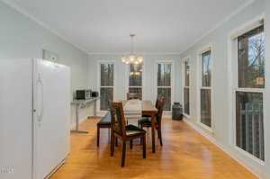 A dining room with a wooden table set for six, surrounded by chairs on a light hardwood floor. Large windows offer a view outside. A white refrigerator and a small black microwave are on the left. Warm overhead lighting creates a cozy ambiance.