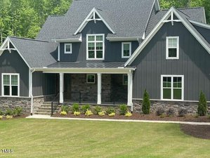 A modern two-story dark gray house with white trims and multiple peaked roofs. The facade features large windows, stone accents, and a covered porch. The well-maintained front yard has grass and small shrubs.