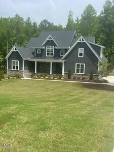 A two-story gray house with white trim and a gabled roof is surrounded by a well-maintained lawn. The front yard features small shrubs and trees. Dense green woods are visible in the background under a clear sky.