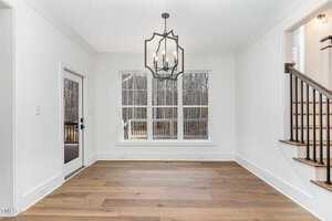 A bright, empty dining room features hardwood floors and a large window with a view of bare trees outside. A black chandelier hangs from the ceiling, and a sliding door to the left opens to a deck. A staircase is partially visible on the right.