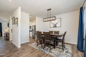 A modern dining room with a wooden table and six chairs on a patterned rug. The walls are white, featuring a framed picture of a longhorn. A contemporary light fixture hangs above the table, and a glimpse of the kitchen is visible in the background.