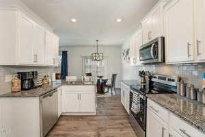A modern kitchen with white cabinets, granite countertops, and stainless steel appliances. The kitchen leads to a dining area with a circular table and blue chairs. The floor is wooden, and a chandelier hangs above the dining table.