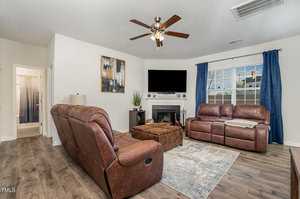 A cozy living room with brown recliner sofas, a patterned rug, a ceiling fan, and a wall-mounted TV above the fireplace. Blue curtains frame the window, and a door leads to a bathroom. A modern painting and potted plant add decor.