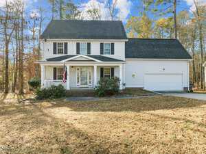 Located at 2786 Clifton Avenue in Creedmoor, this charming two-story white house features a welcoming front porch, black shutters, and a double garage. The American flag waves proudly near the entrance. Surrounded by trees, it rests under a blue sky with scattered clouds while the lawn is mostly brown.