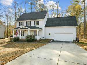 A two-story white house with black shutters and a dark roof is surrounded by trees. An American flag hangs on the porch. There's a double garage and a concrete driveway in the foreground. The season appears to be fall.