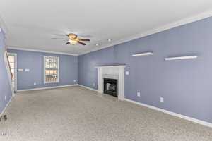 A spacious, empty living room with light blue walls, white trim, and carpeted floor. It features a ceiling fan with lights, a white fireplace, and two small white shelves on the wall. Natural light enters through a window on the left.