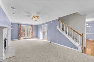 Spacious living room with light blue walls and carpeted floors. A ceiling fan is centered above. French doors with floral curtains lead to an outdoor area. A staircase with a wooden railing ascends to the right, and a white fireplace is on the left.