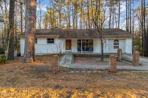 A single-story white house on 404 E D Street in Butner boasts a brown shingle roof and is surrounded by tall trees. The front features a covered porch and a brick-paved area. A curved driveway leads to a red brick entryway framed by two brick pillars.
