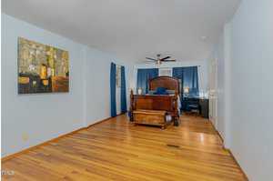 A spacious bedroom with hardwood floors and light blue walls. It features a wooden bed with blue bedding, a chest at the foot, a painting on the left wall, dark blue curtains, and a ceiling fan.